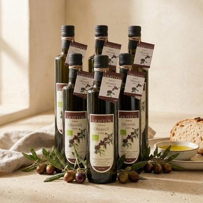 Sechs Flaschen ARISTOS Natives Olivenöl Extra 500 ml gestapelt, daneben Olivenzweige, Brot und ein Ölschälchen
